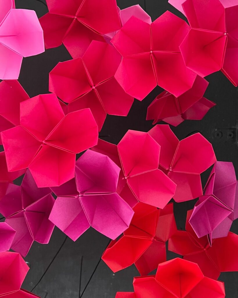 Composition florale en origami réalisée en papier japonais, composée de fleurs aux nuances de fuchsia, mauve et rouge sur une structure en bois noir et des tiges métalliques.